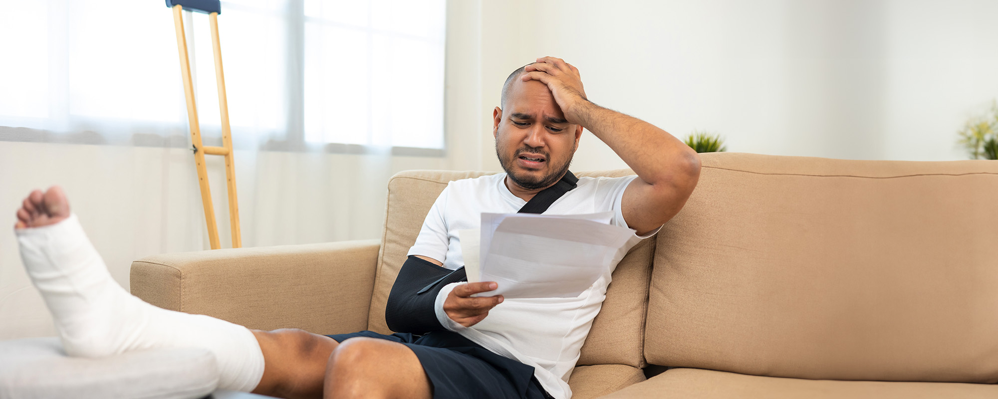 Man on couch with leg in cast elevated, his arm in sling grasping his head in pain. Orange County Injury Lawyers concept image
